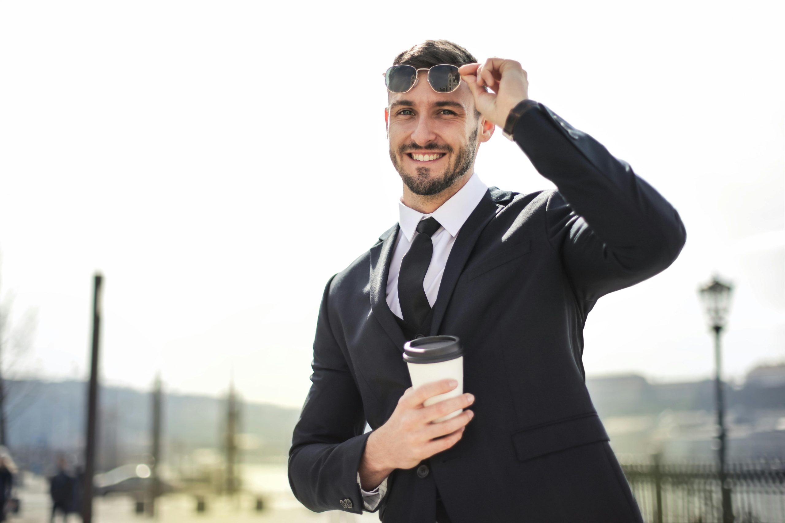 Smiling businessman in formal attire with coffee cup enjoying a sunny day outdoors.